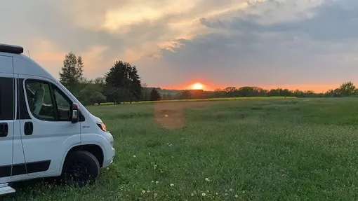 Ein weißer Kastenwagen-Camper steht am Rand einer grünen Wiese bei einem malerischen, orange-roten Sonnenuntergang auf dem Wohnmobilstellplatz des Geflügelhof Arnsdorf.